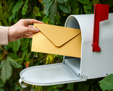 putting mail in white mailbox with red flag and greenery in the background