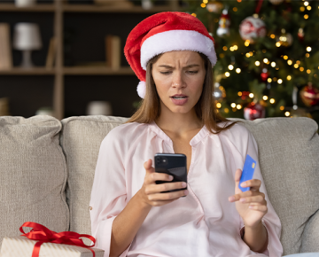 woman sitting on couch in Santa hat viewing phone and credit card with confused look