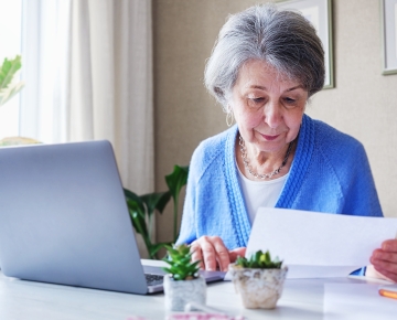elderly woman in blue looking at paper next to laptop