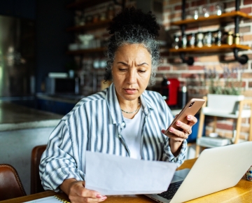 woman at laptop holding phone and looking at paper